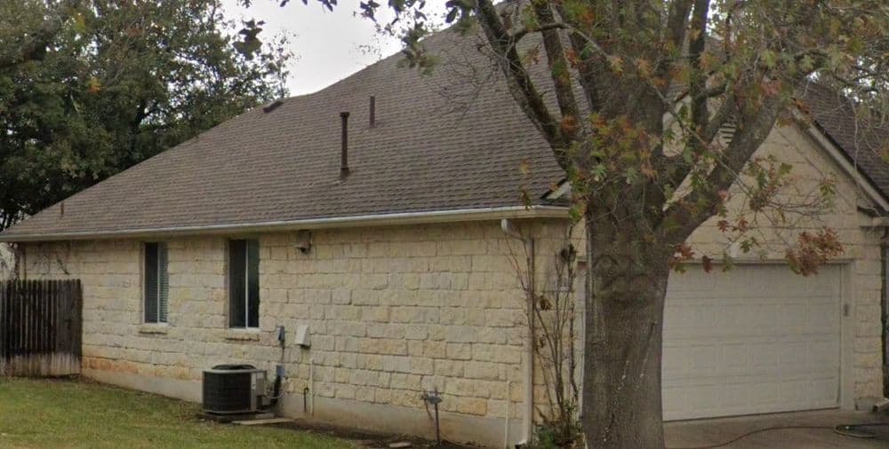 Single-story stone house with garage, surrounded by trees and grassy area.