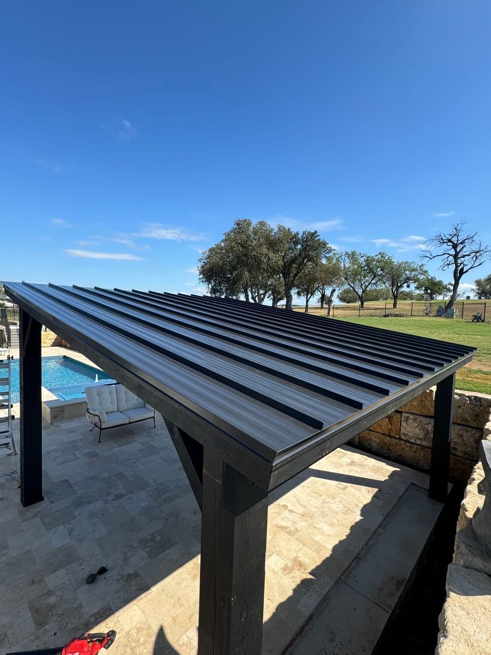 Modern metal roof pergola over patio by pool, surrounded by trees and blue sky.