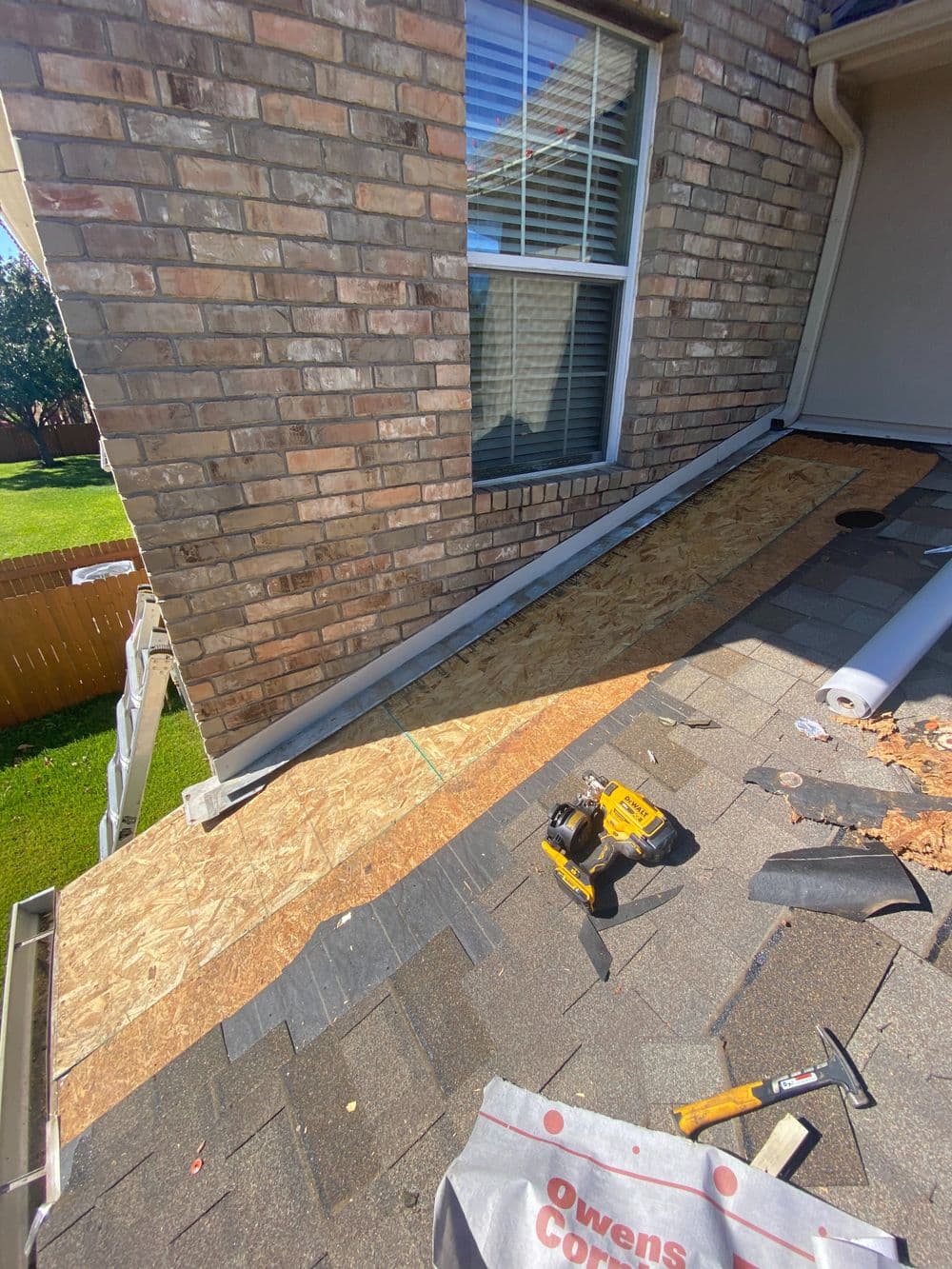 Roof renovation work with plywood decking, tools, and shingles near a brick wall and window.