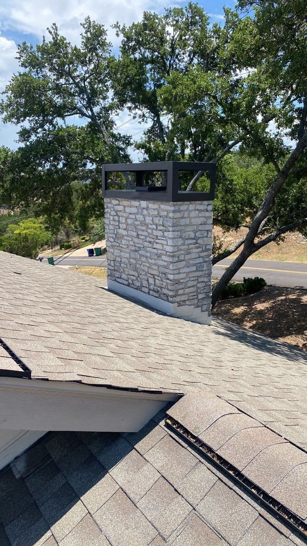 Chimney with stone facade and metal cap atop a sloped roof, surrounded by trees.