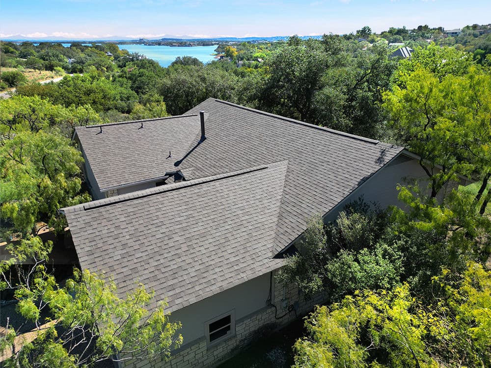 Aerial view of a modern home with a dark roof surrounded by greenery and water in the distance.