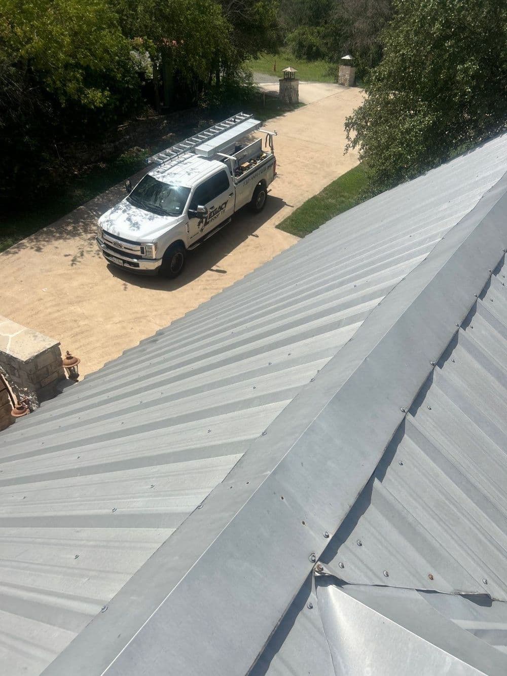 Metal roof view with a service truck parked on a driveway surrounded by greenery.