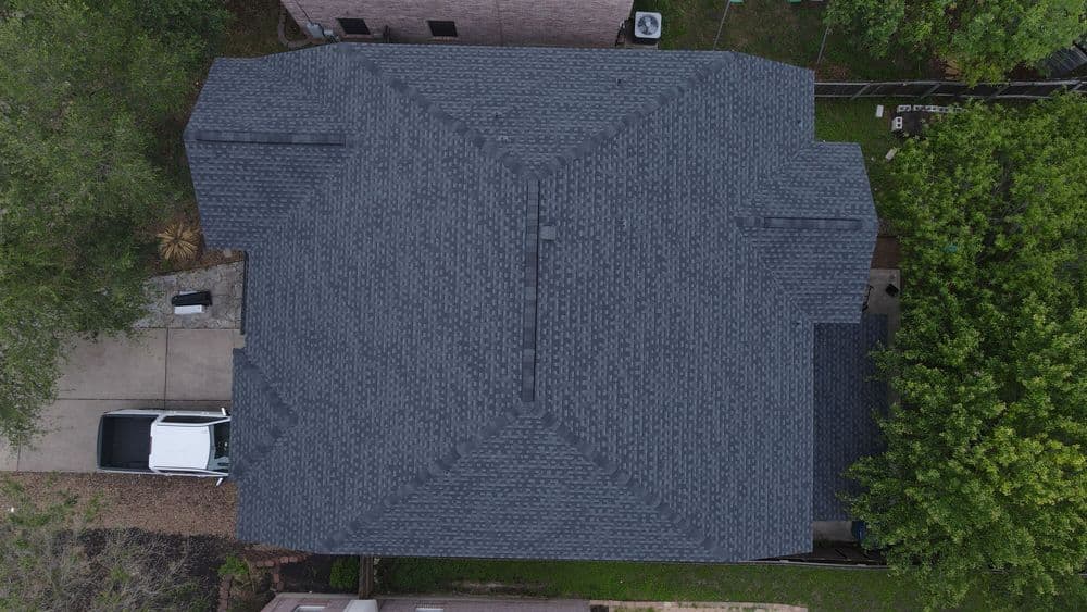 Aerial view of a gray shingled roof with surrounding trees and a parked vehicle.
